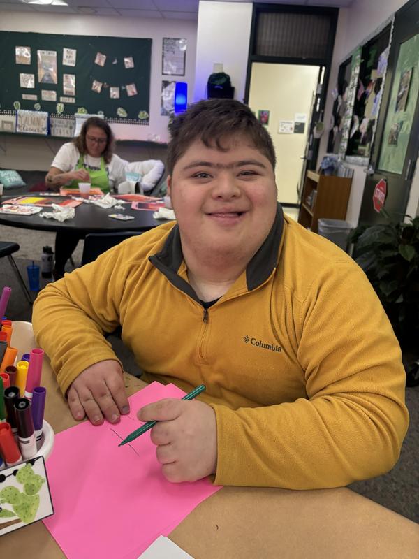 A boy smiles while drawing on pink paper at a table with art supplies.