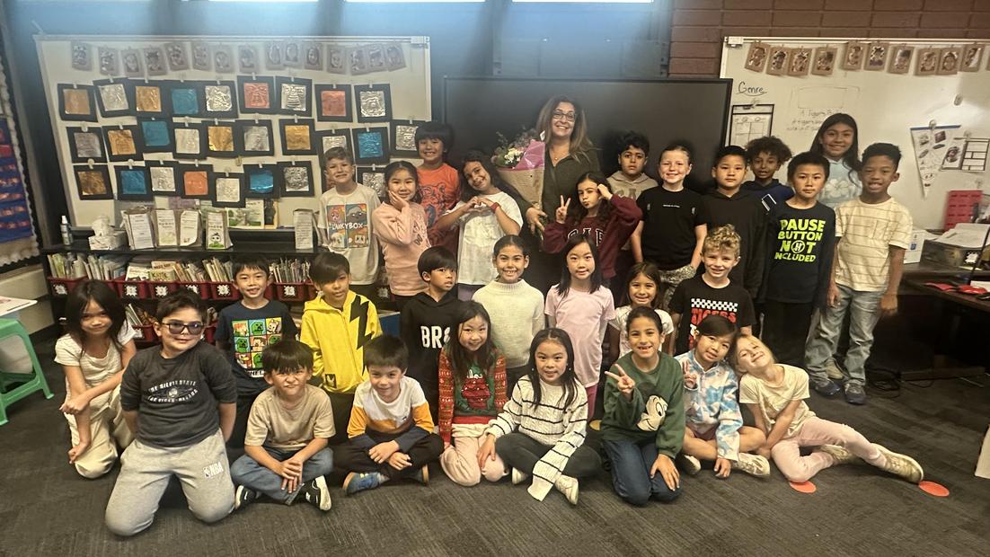 A large group of smiling students posing in a classroom with a teacher holding flowers.