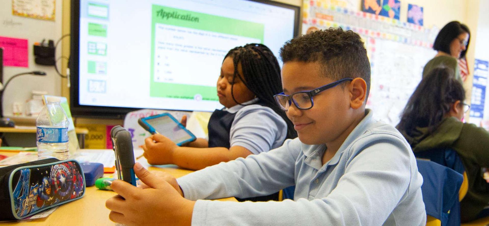 Students engaged with devices at a classroom table, one using a smartphone.