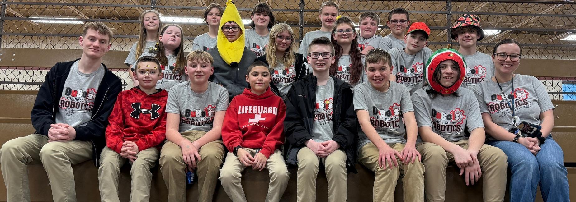 A large group of students in matching t-shirts poses for a photo at a robotics event.