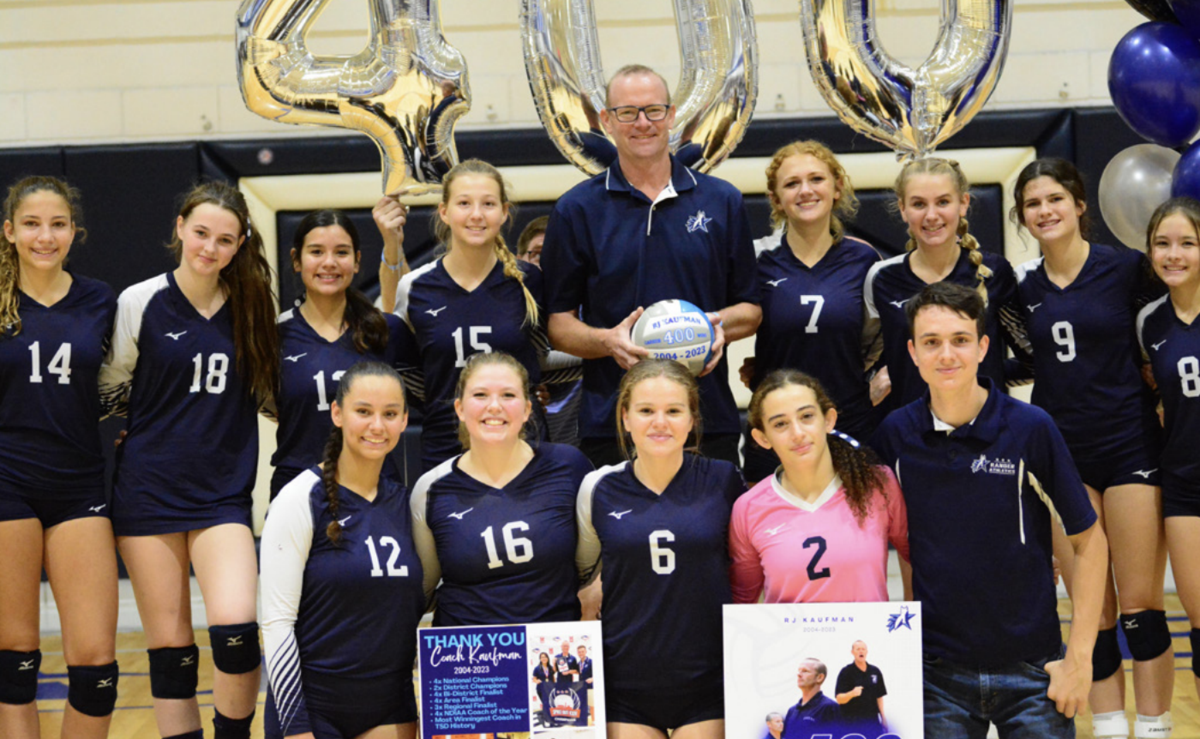 Group photo of a volleyball team and Coach RJ Kaufman celebrating his 400th career win. The coach stands at the center, holding a volleyball marked with ‘400 – 2023,’ while two players behind him hold large silver ‘400’ balloons. The team members are dressed in navy and white uniforms, with one player in a pink jersey. They are all smiling and showing appreciation for the coach, who stands proudly among them. Two team members in front hold a poster thanking the coach for his contributions.