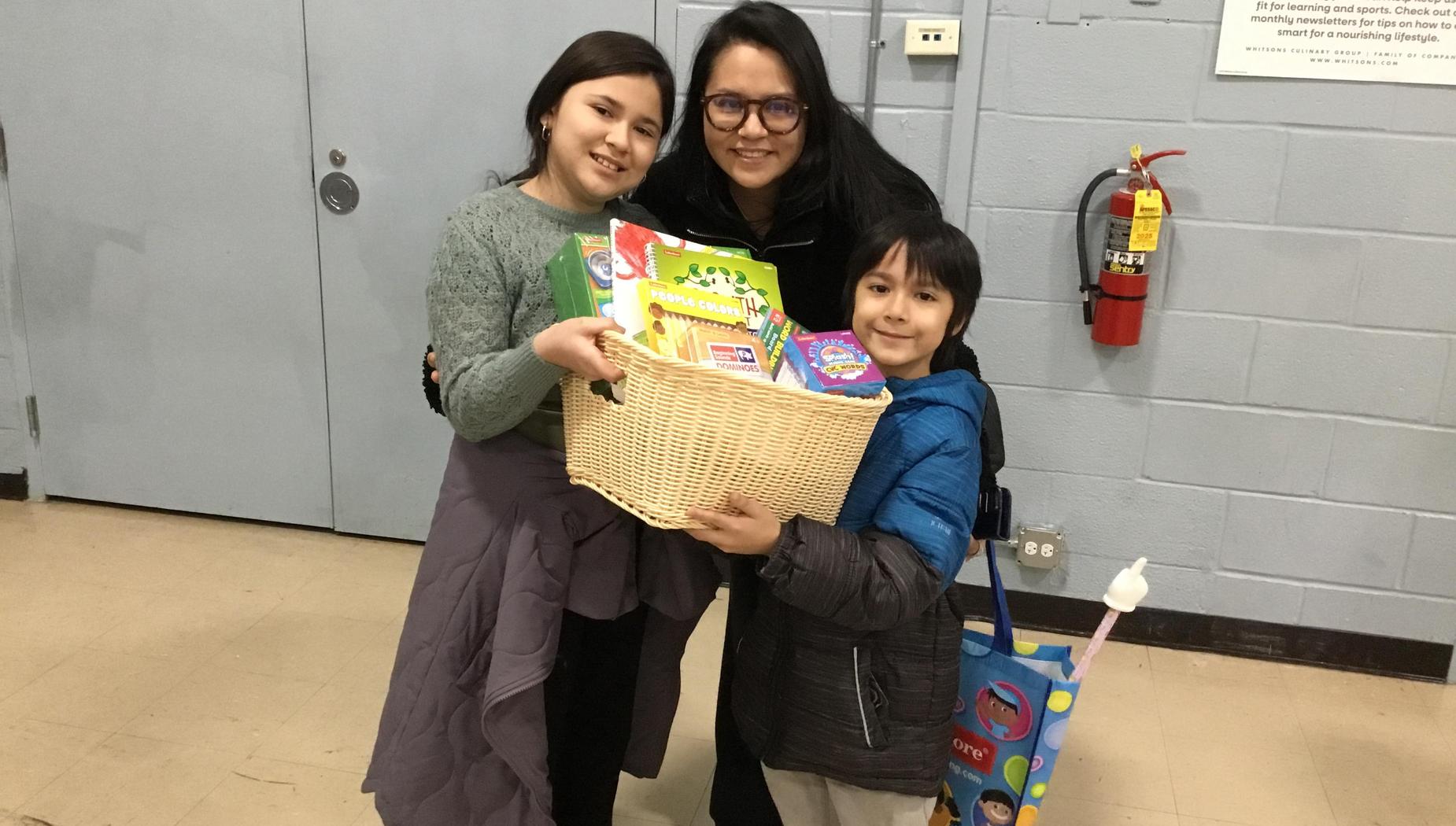 Two children and an adult pose together with a basket of toys in a hallway.