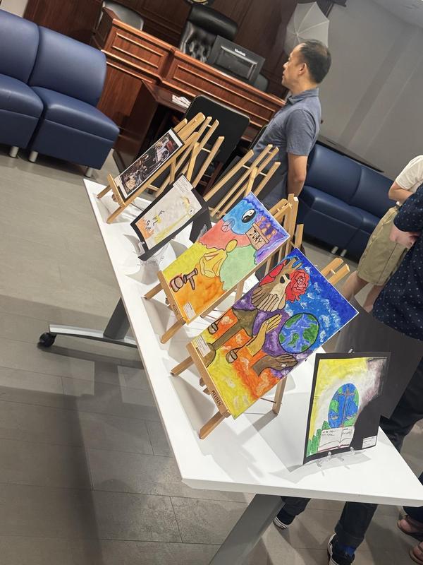 Display of colorful artworks on easels with visitors observing in a courtroom-like setting.