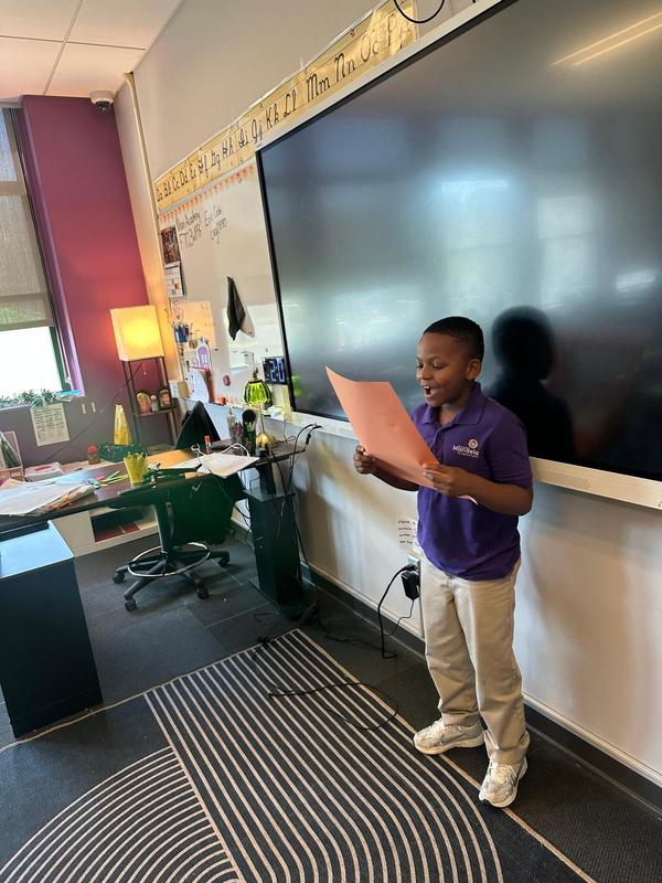 A scholar stands at the front of the classroom reading from an orange piece of paper.