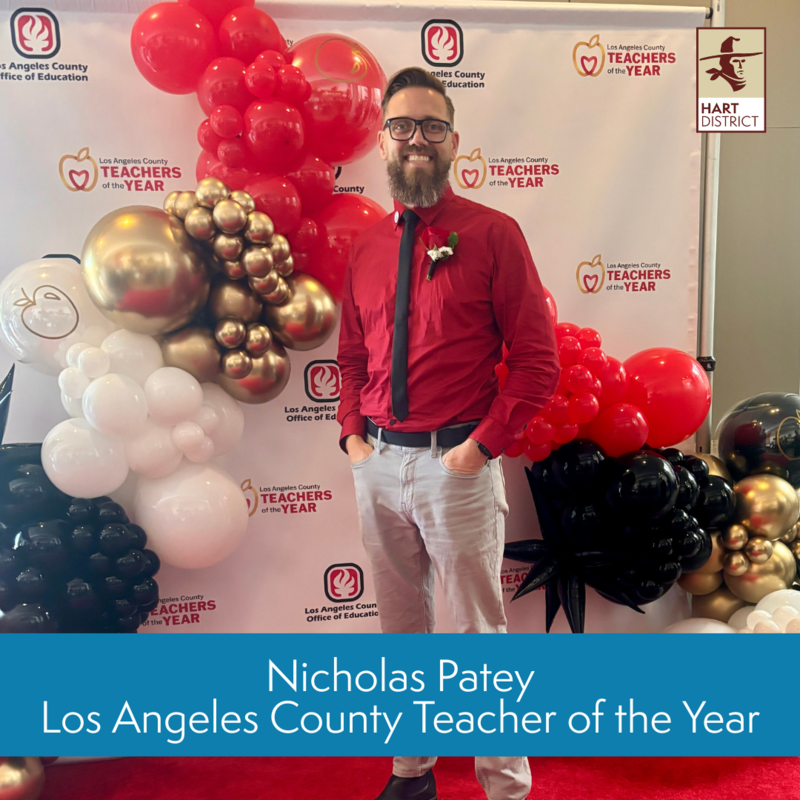 Nicholas Patey, a bearded man with glasses, poses in front of the Los Angeles County Office of Education Teacher of the Year banner with red and gold balloons.