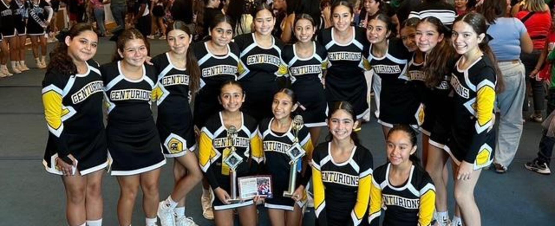 Cheer team with trophies posing in a large crowded hall.