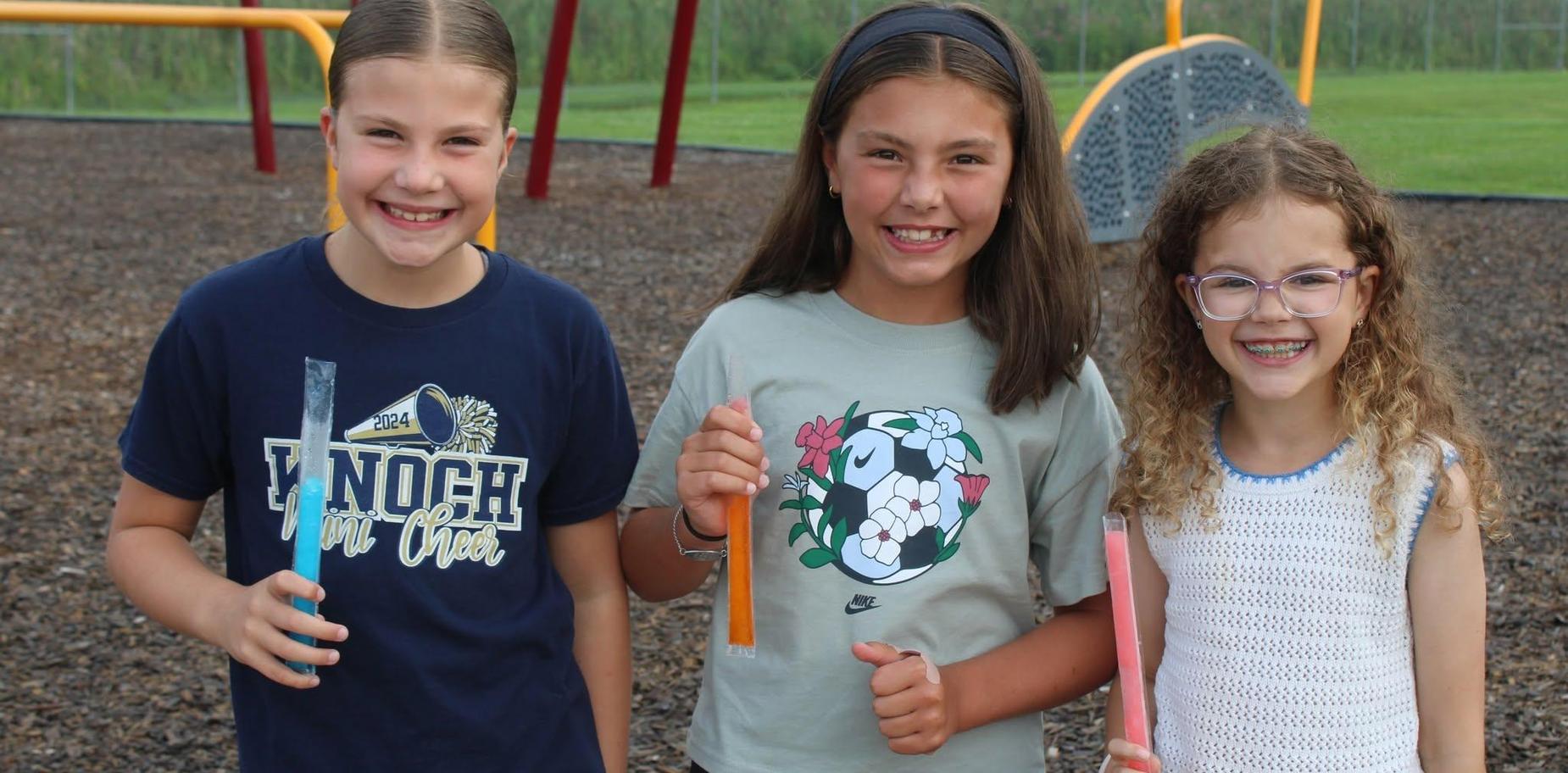 Three girls smile while holding colorful freeze pops at a playground.