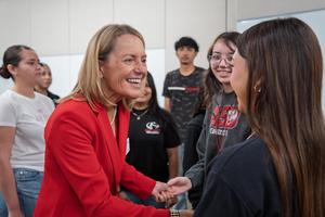 A smiling woman in a red blazer shakes hands with a student while others look on in the background.