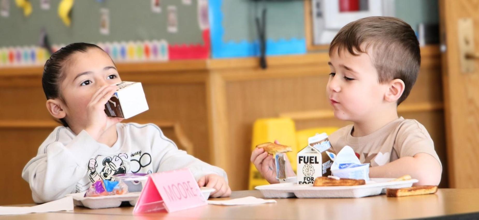 Two children eating lunch at a table with milk and snacks.