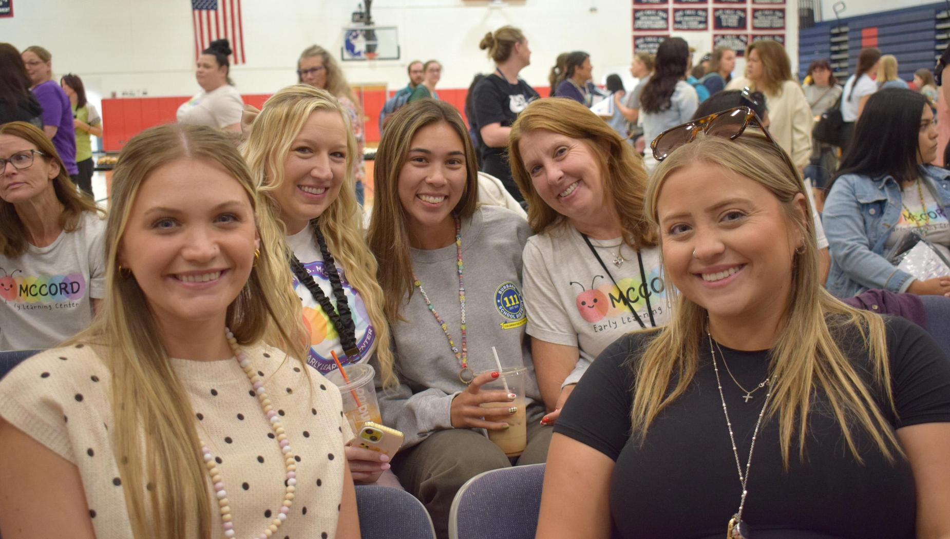 Five smiling women sitting together, all wearing casual attire with drinks.