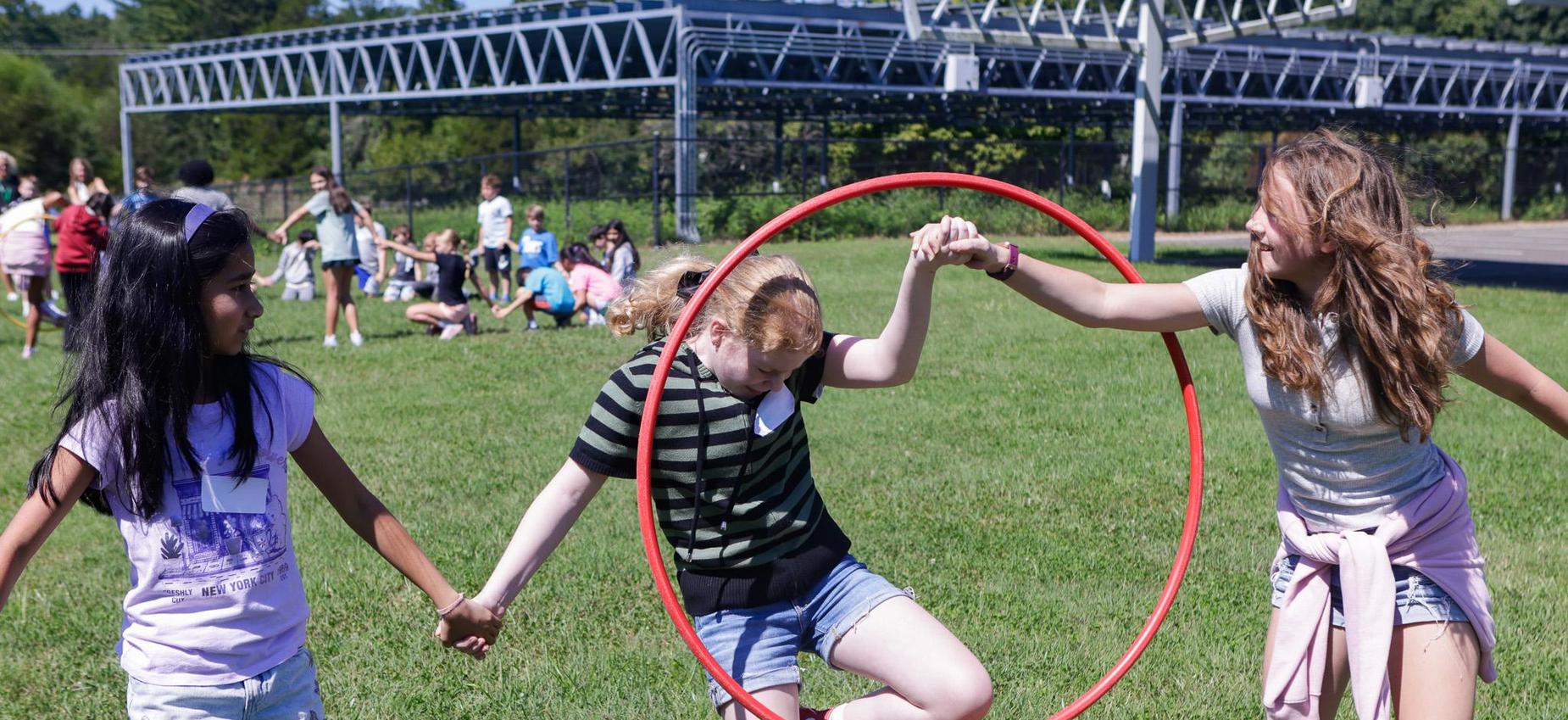 Three children play outdoors, holding hands and interacting with a hula hoop.
