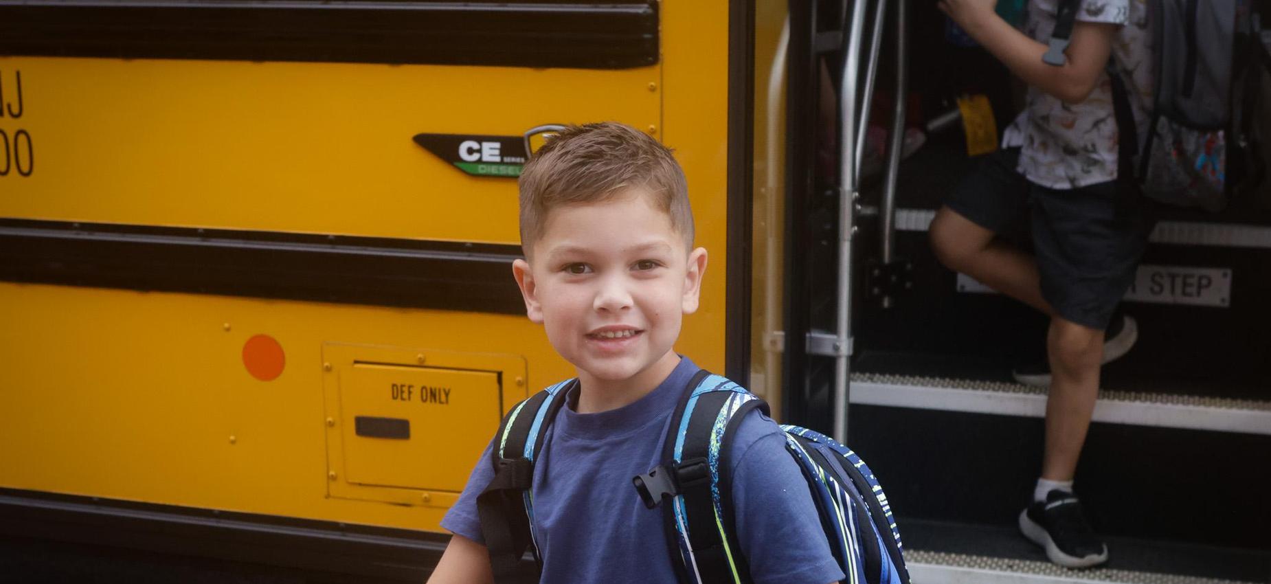 Smiling boy with a backpack stands at a school bus entrance.