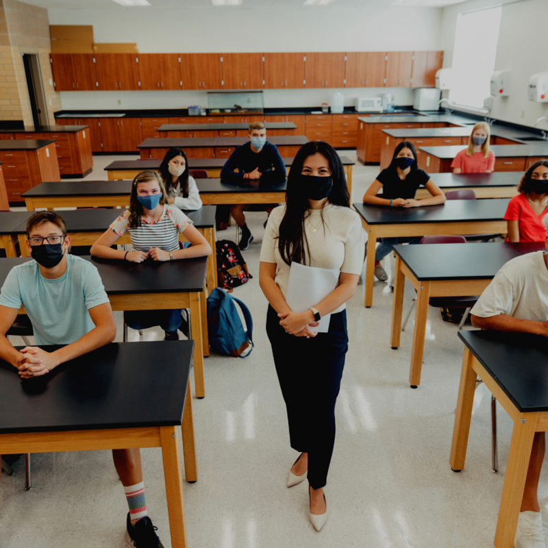 students seated at tables in facemasks and teacher standing in center in facemask