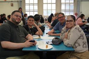 Four people smiling at a table with food in a busy room.