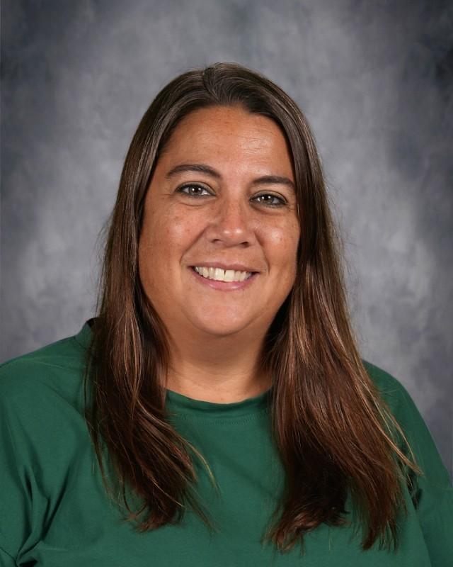 Smiling woman with long brown hair wearing a gray shirt with colorful 'TEACHER' text.