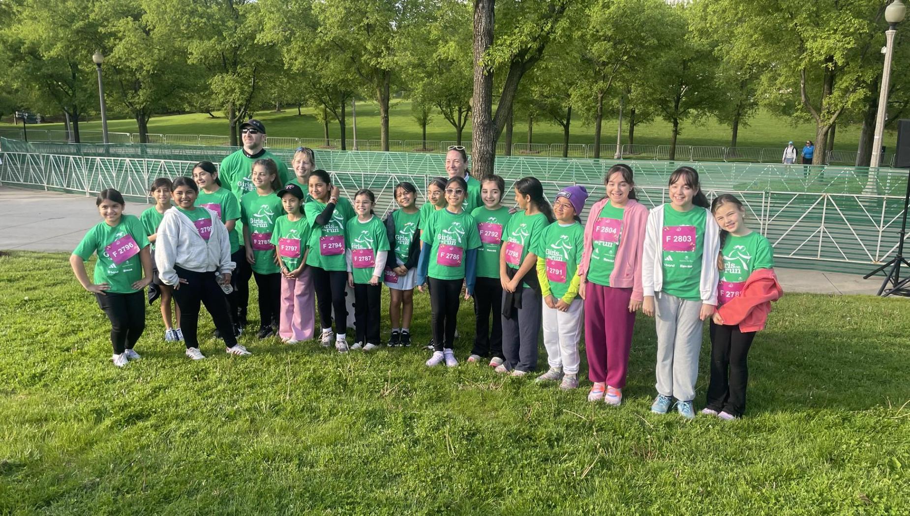 Group of children in green shirts smiling and standing on grass outdoors.