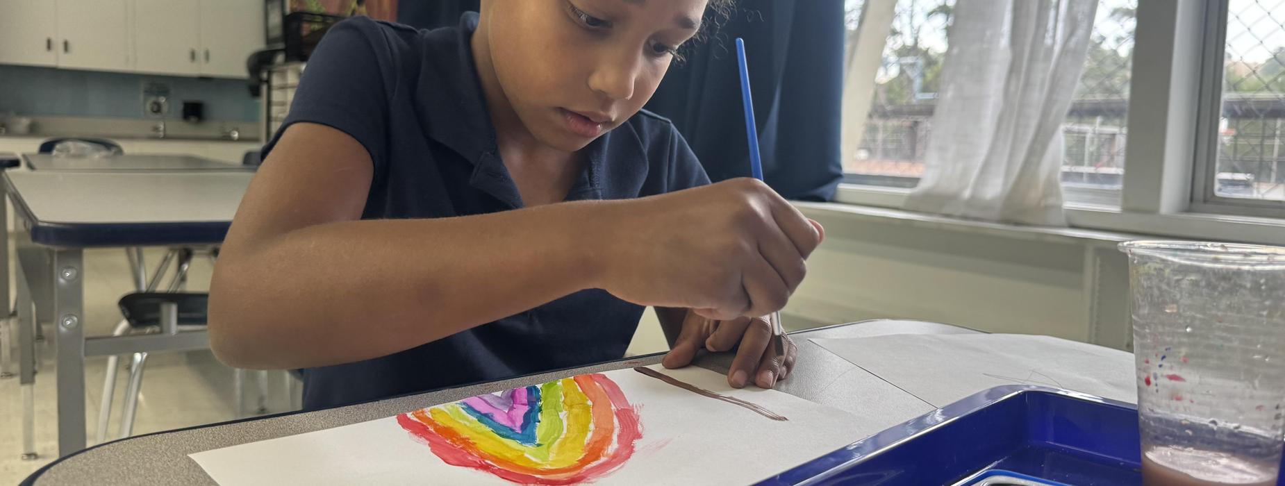 A girl focused on painting a rainbow with a brush on paper at a classroom table.