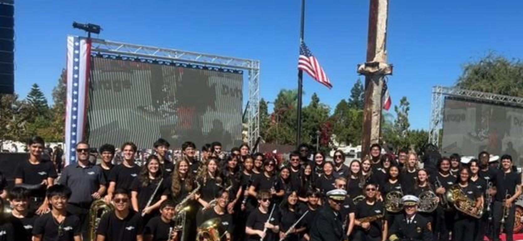 A large group of musicians poses together outdoors with flags and screens behind them.