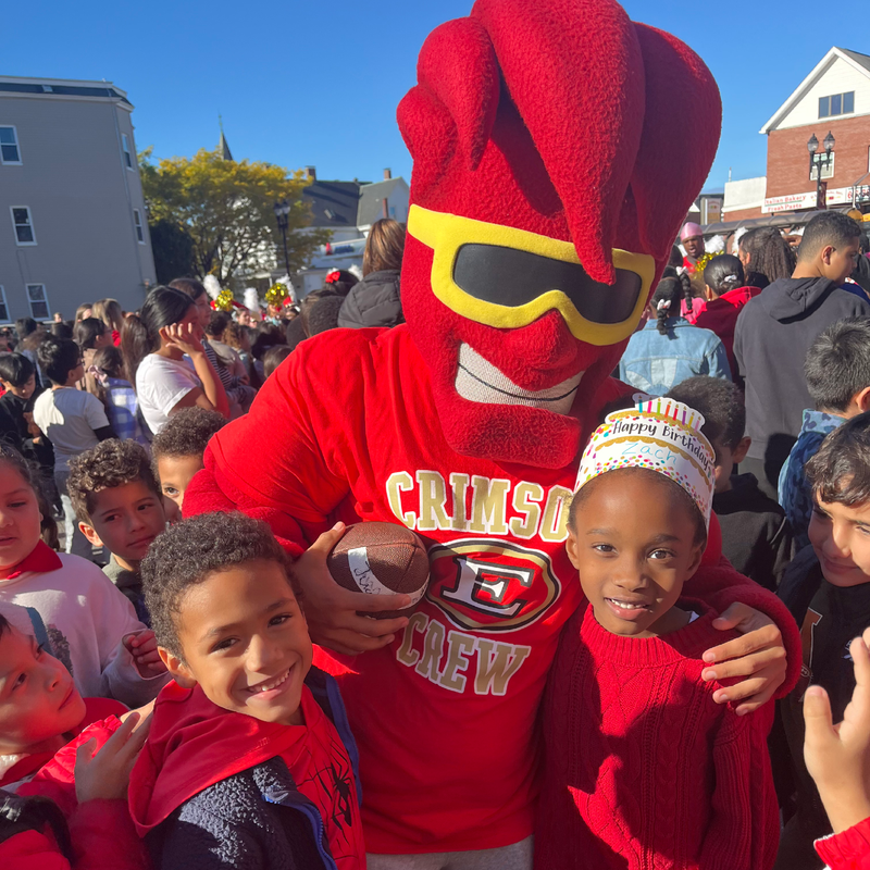 A red mascot in sunglasses holds a football, posing with two smiling children at a crowded event.