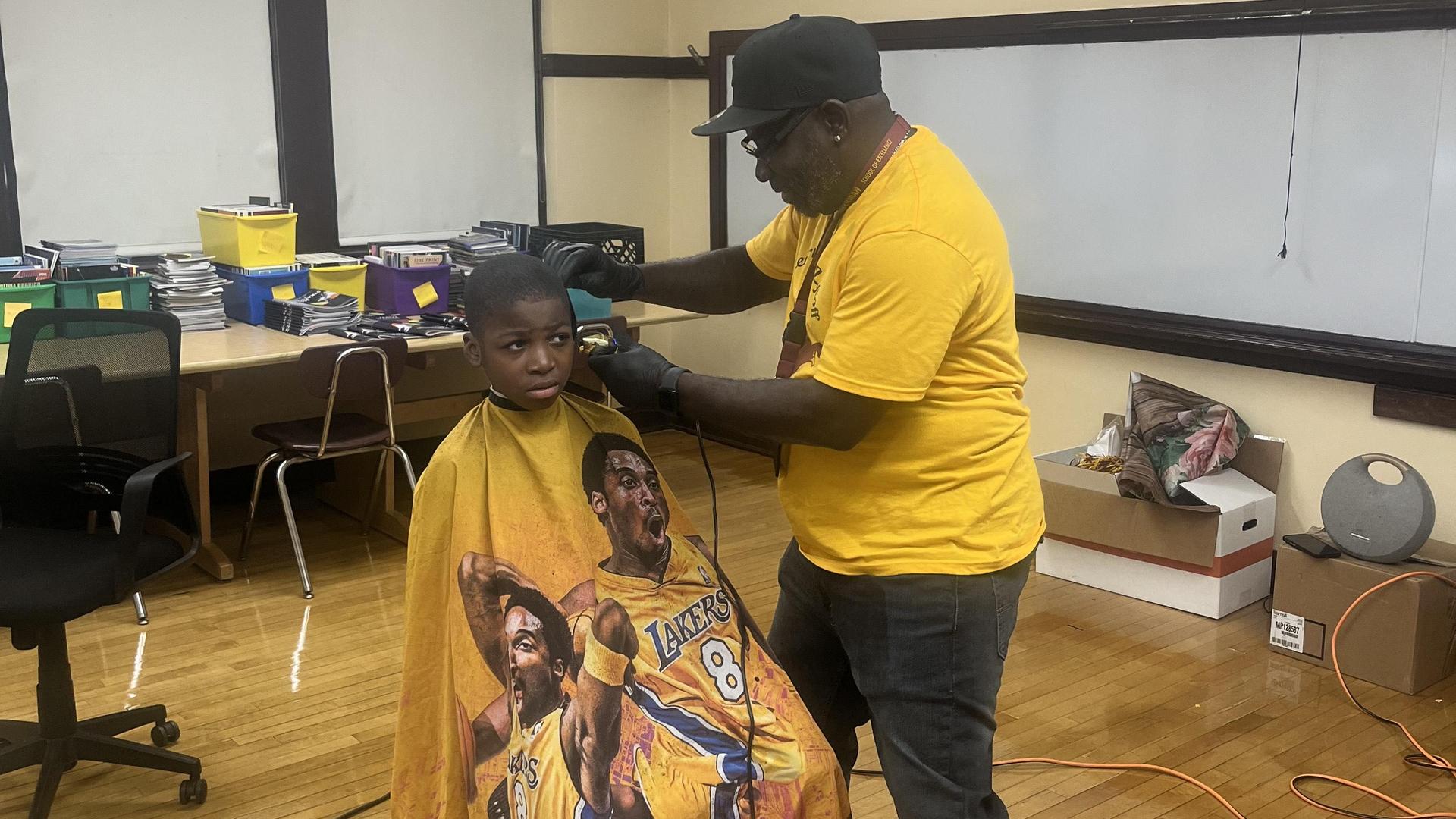 A boy in a barber's chair receiving a haircut while wearing a Lakers-themed cape.