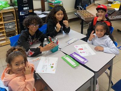 Group of children sitting at a table engaged in activities, smiling and showing hand signs.