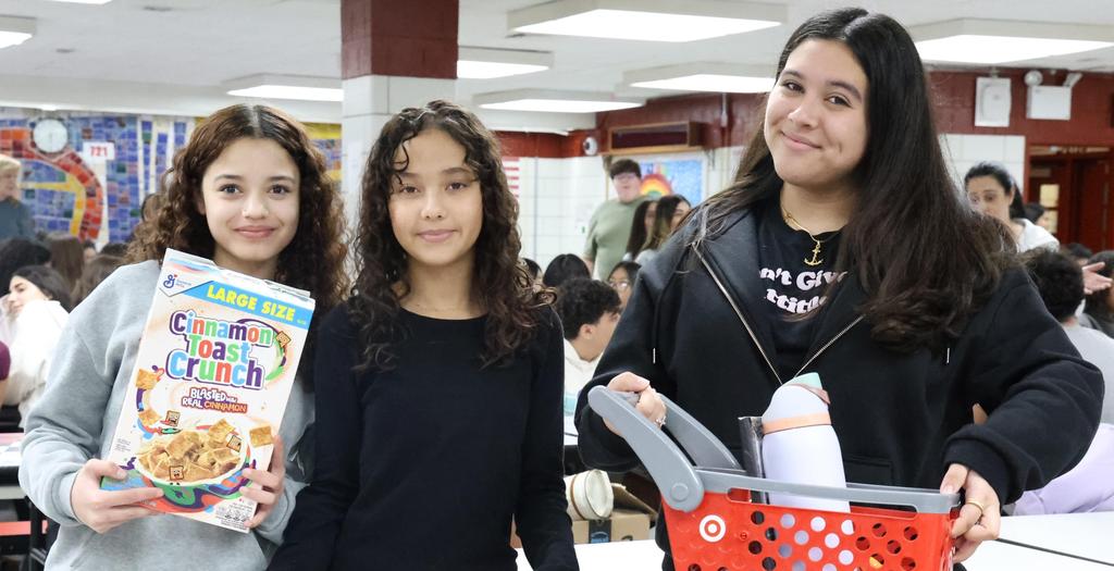 Three girls standing.  One girl is holding a cereal box. One girl is holding a pink crate. The third girl is holding a miniature red shopping cart.