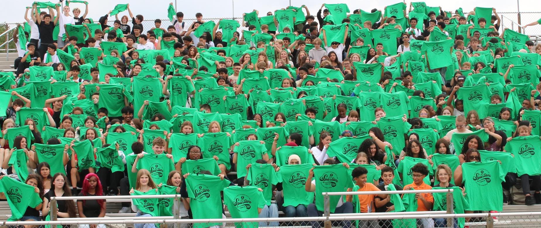 Students holding up green shirts in bleachers.