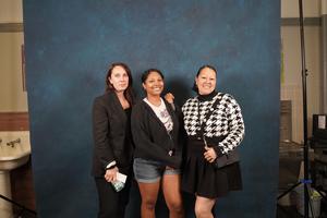 Three women posing together in front of a blue backdrop, dressed stylishly.