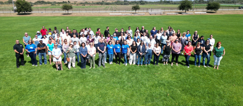 A large group of diverse people standing together on a grassy field with a clear sky.