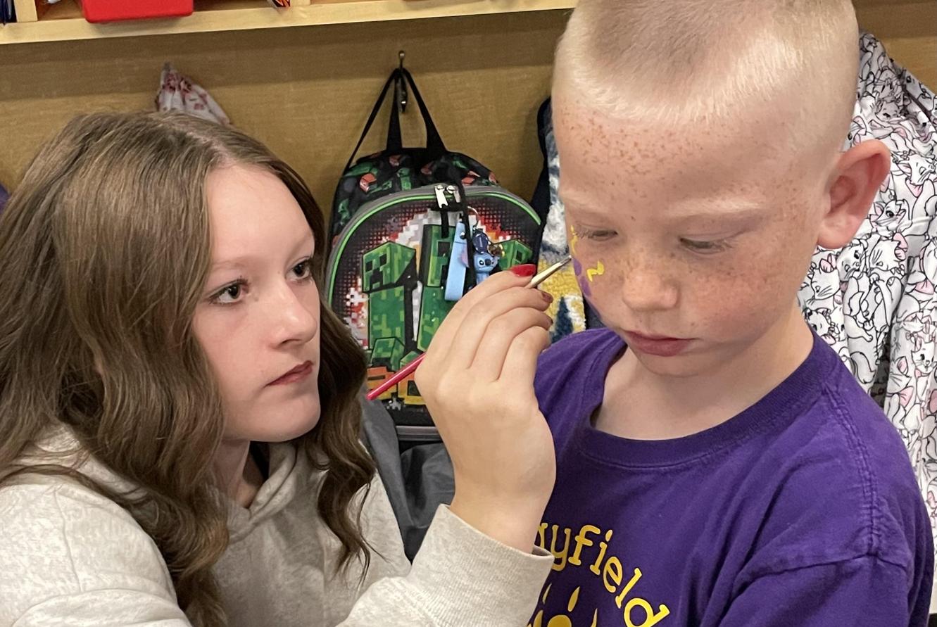 A girl painting a design on a boy's face while he watches intently.