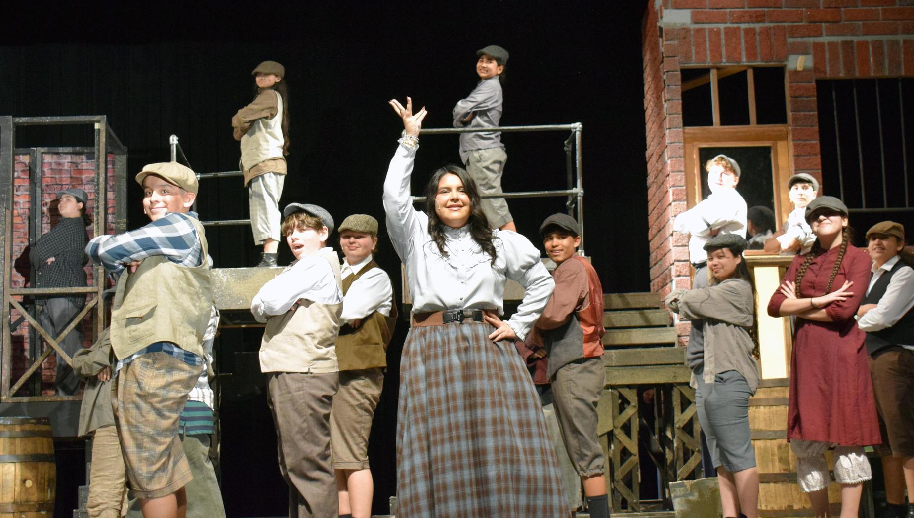 A large ensemble of actors in vintage clothing posing on stage with props.