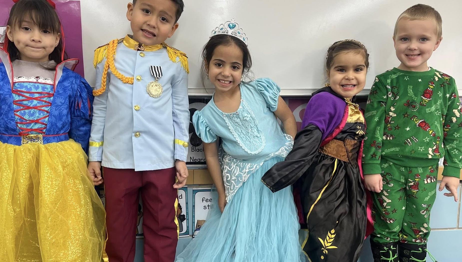 Five children in colorful costumes smiling together in a classroom setting.