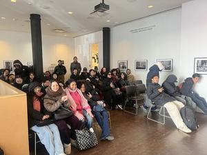 A large group of people sits and poses for a photo in an art space with black and white photographs.