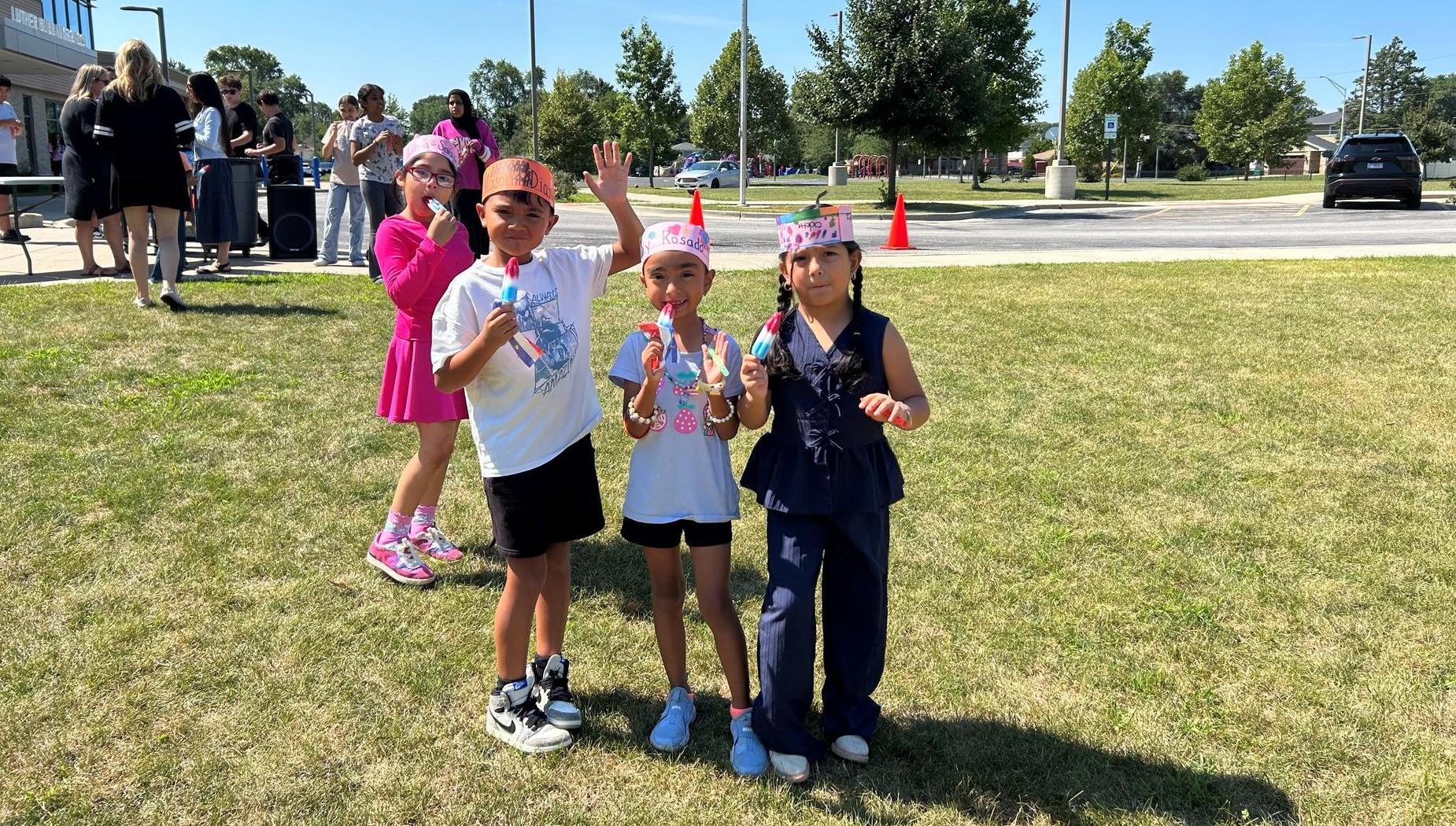 Three girls holding ice pops standing on a grass field.