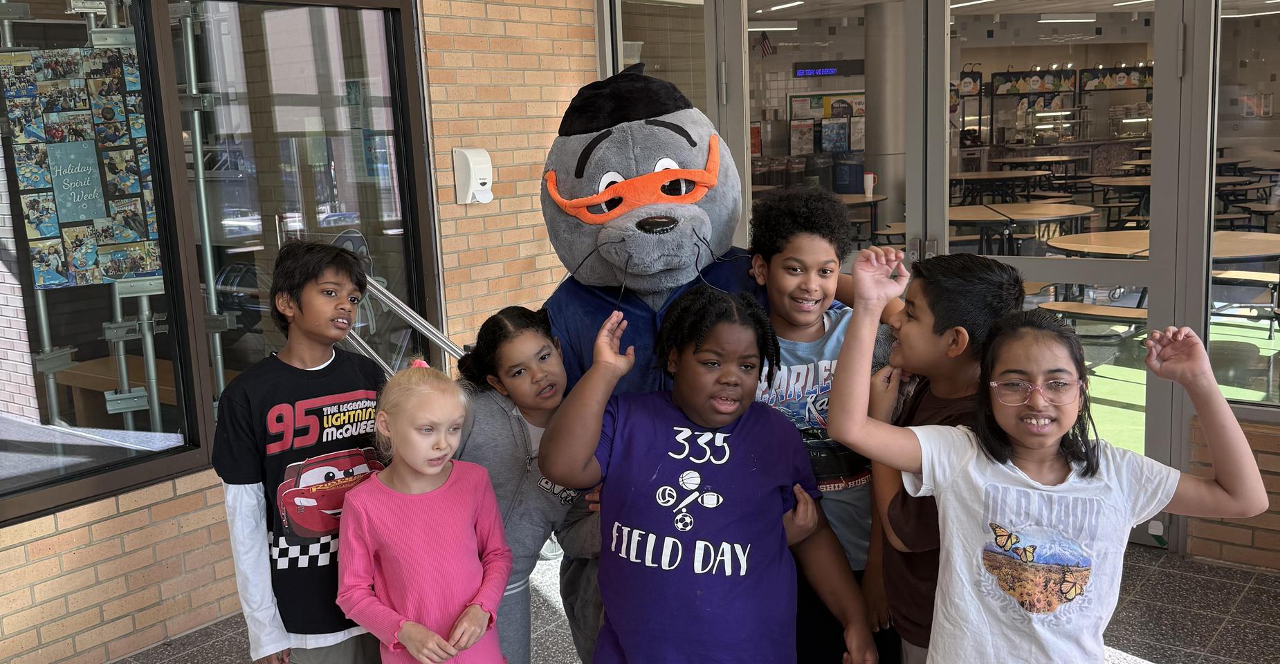 A mascot stands among a group of children in a school hall.
