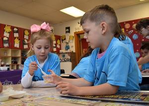 Two children work together, with one using a spoon to mix in a small cup at a table.