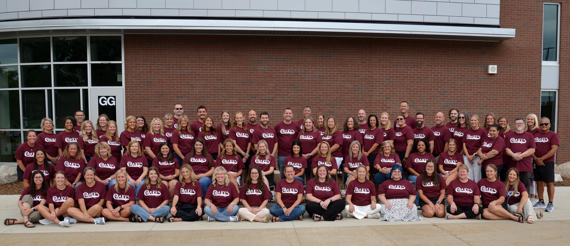 the staff wears their maroon shirts with the word Oakes on the front while posing in three rows outside the front of the school