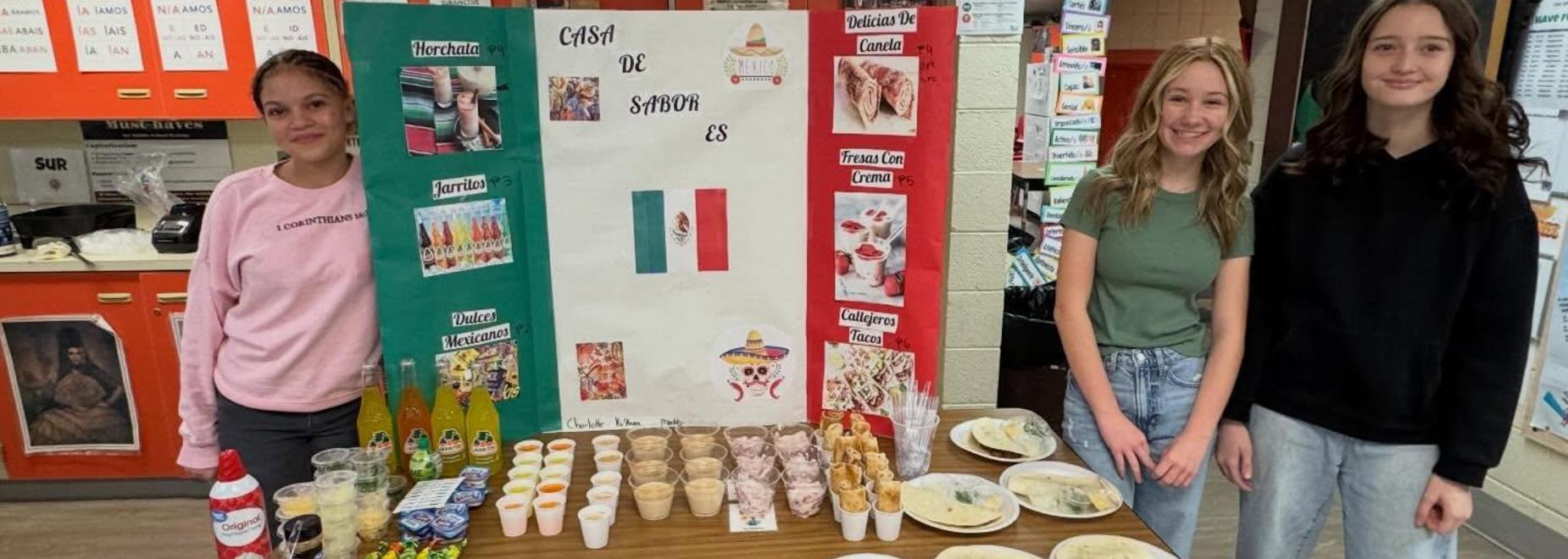 Three women stand beside a decorated table of food and drinks for a cultural presentation.