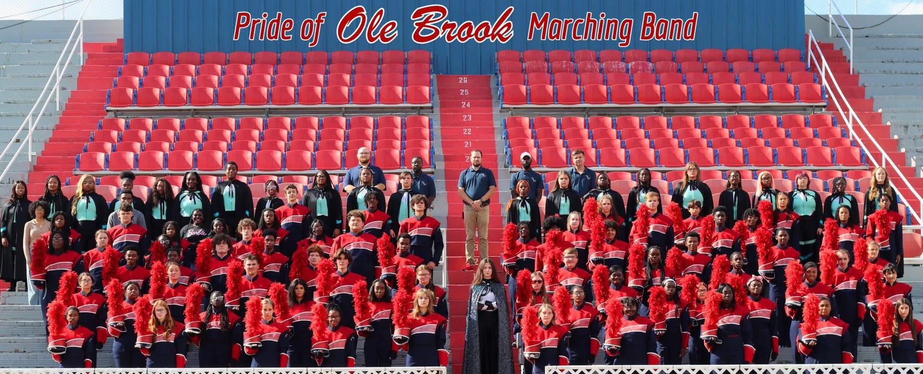 Marching band members in red and blue uniforms pose on stadium bleachers.