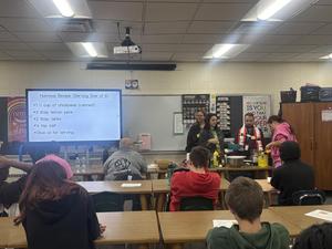 Cooking class participants prepare hummus with a recipe displayed on the screen.