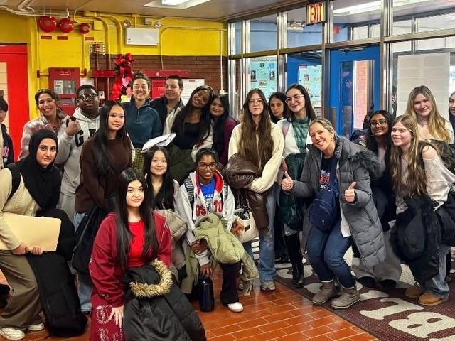 Group of students of diverse backgrounds posing indoors, showing thumbs up.