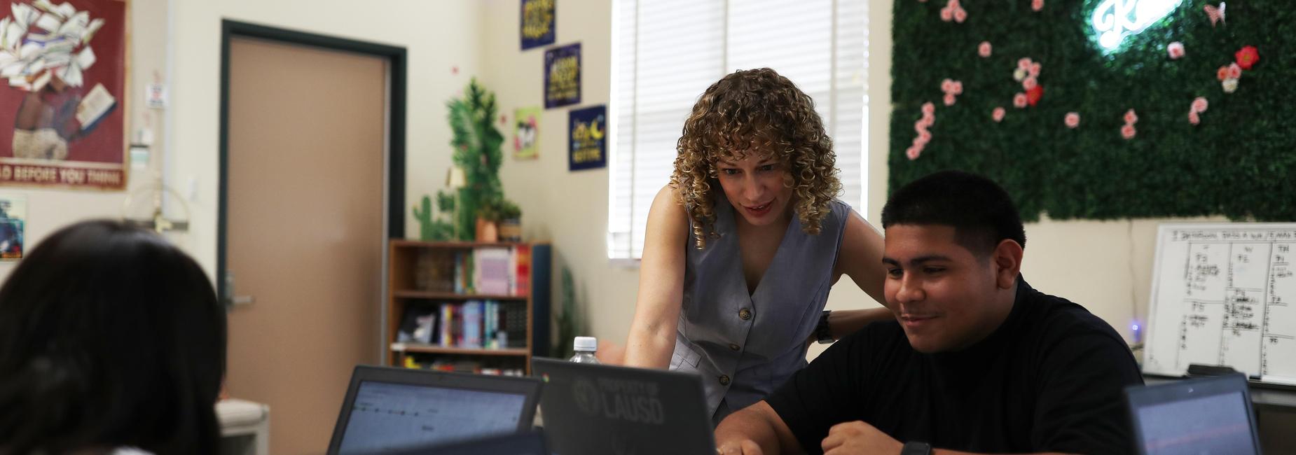 Teacher assisting students with laptops in a classroom setting.