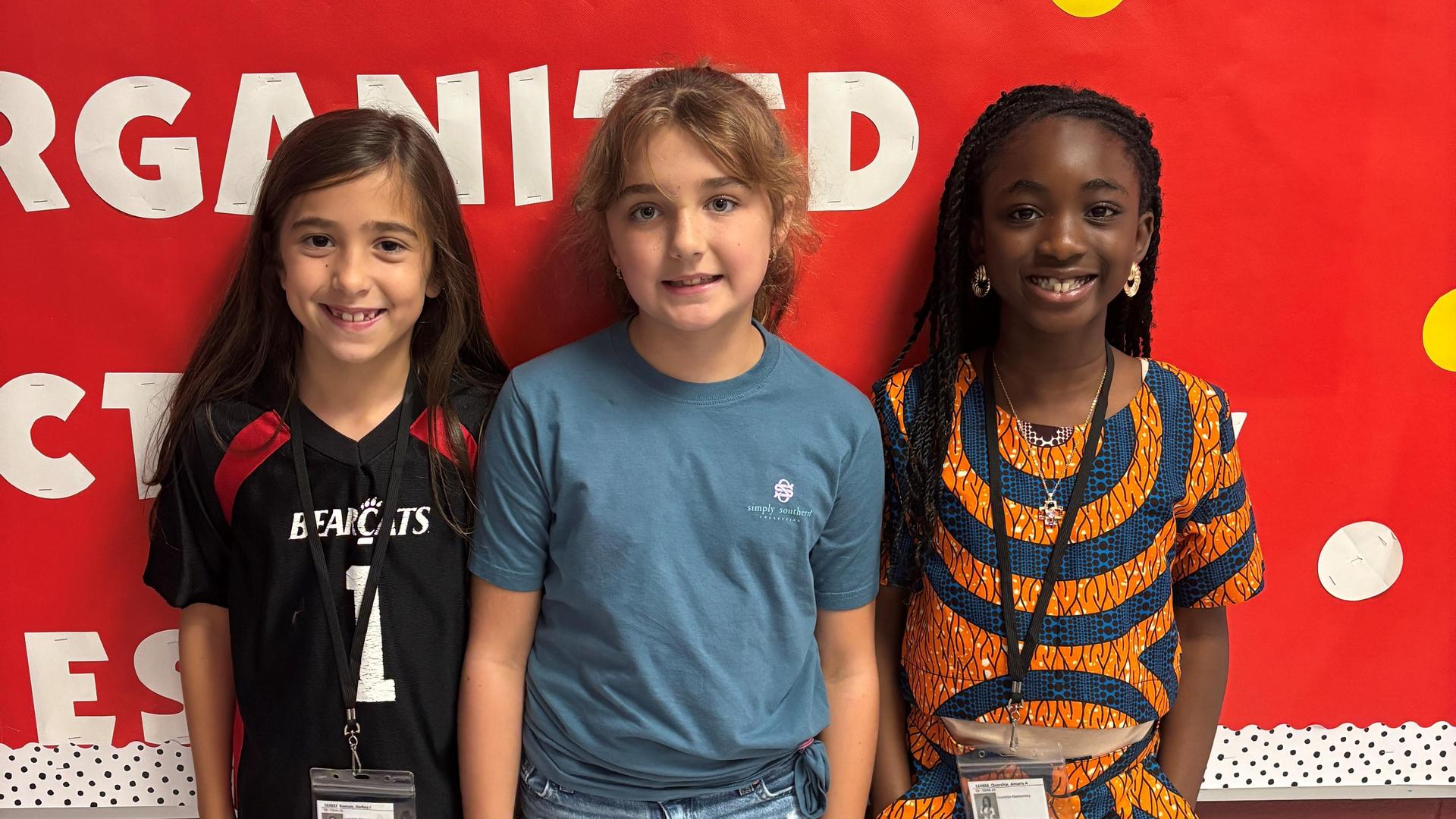 Three girls standing together, smiling in front of a colorful bulletin board.