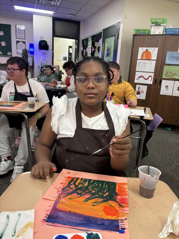 Young girl holding a paintbrush and working on a colorful painting at her desk.
