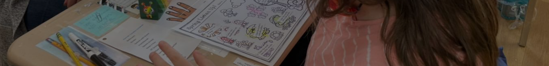 A girl colors a worksheet filled with animals and a handprint art piece on a desk.