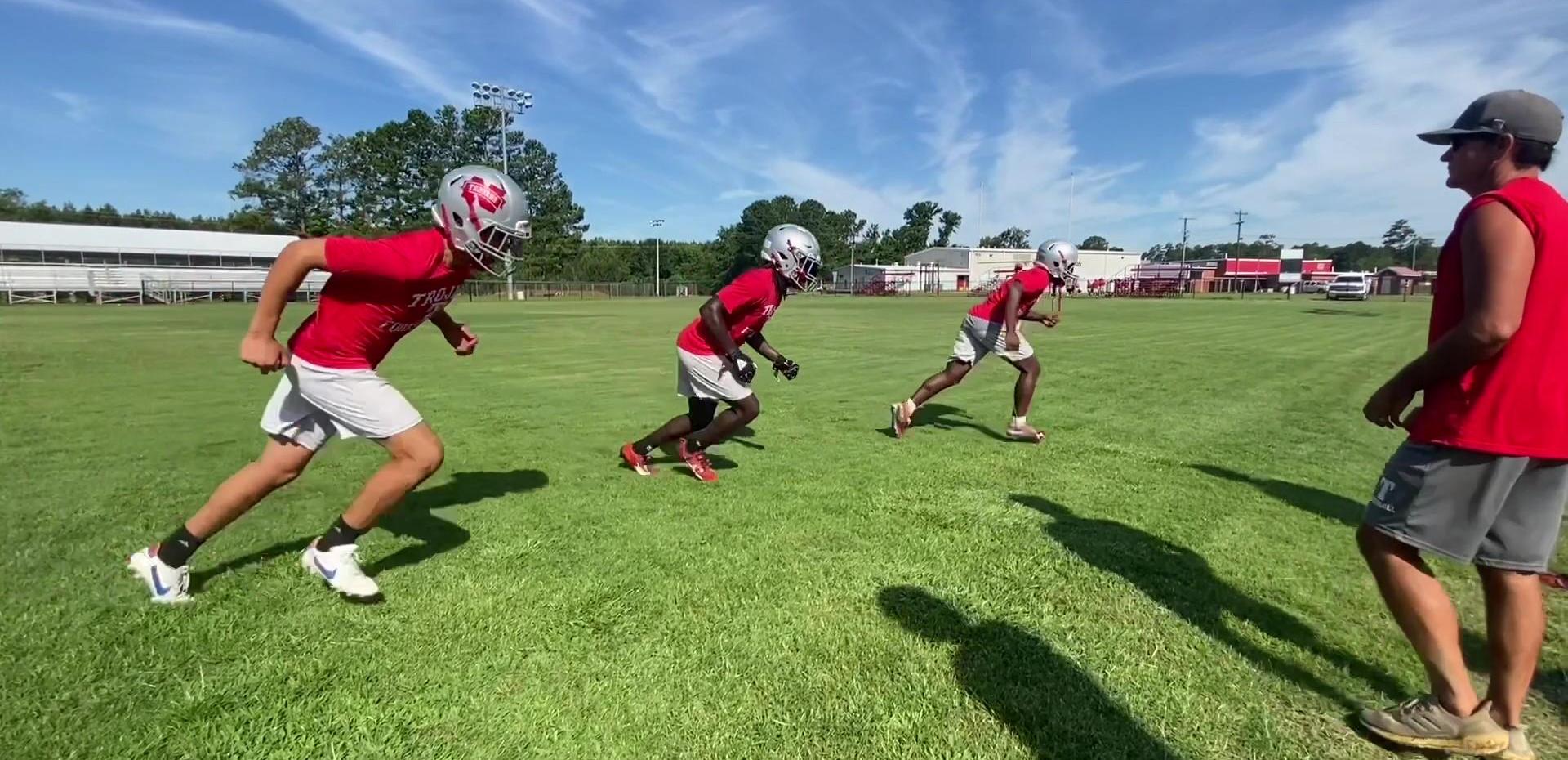 Three football players in red jerseys running on a grass field under a blue sky.