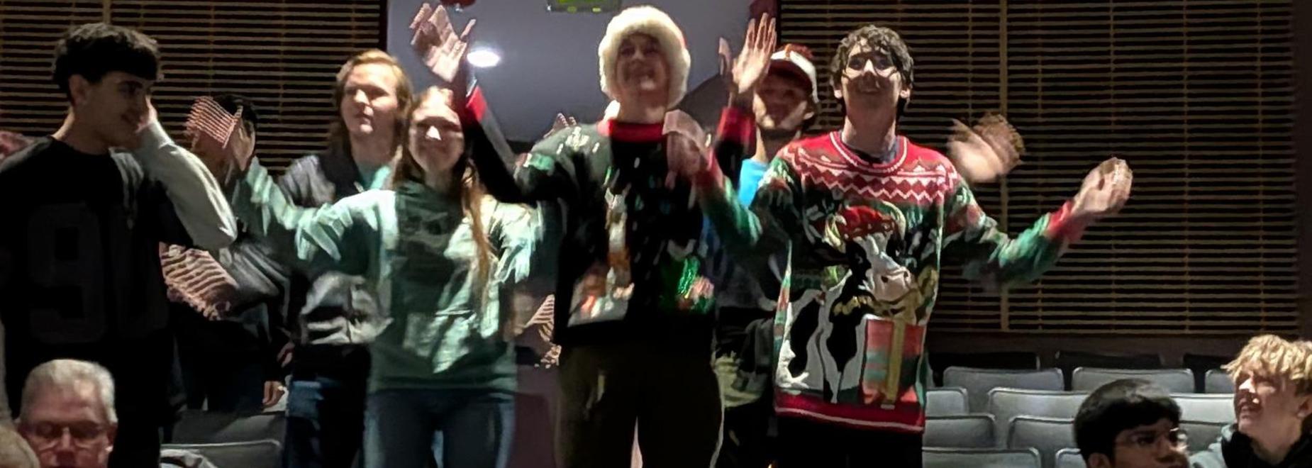 A group of students wearing Christmas sweaters dances joyfully in a theater.