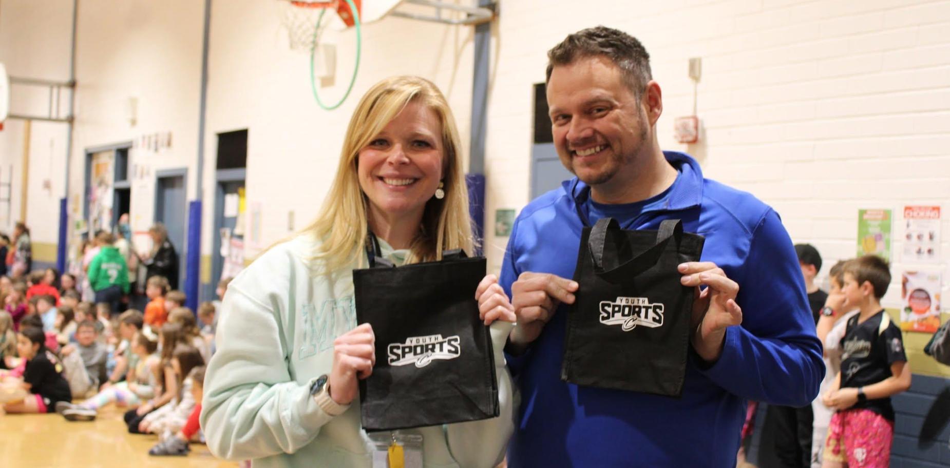 Two adults holding black sports bags in a gym with children in the background.