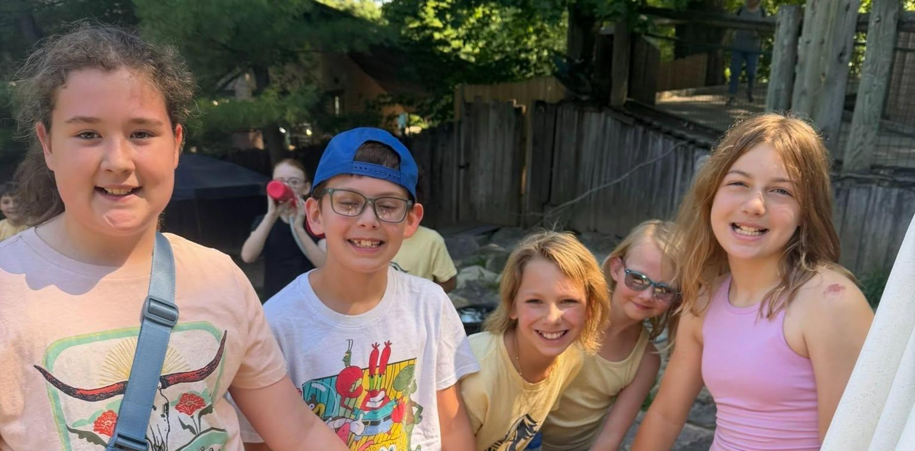 Five children smiling and touching a goat outdoors in a casual setting.