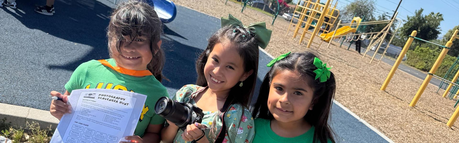 Three children at a playground, one holding a camera and another a photography sheet.